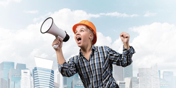 Expressive woman in safety helmet shouting into megaphone. Portrait of young emotional construction worker with loudspeaker on background of modern city. News announcement and advertisement.