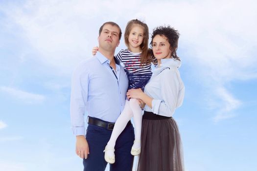 Happy young family, mom dad and little daughter.Parents keep the girl in her arms , and she hugs their neck.On the background of summer blue sky and fluffy clouds.