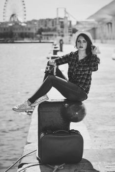 Young woman taking photographs with a vintage camera sitting in a harbour. Girl wearing plaid shirt, blue jeans and sun hat. Black and white photograph.