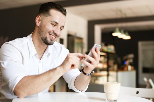 Young caucasian businessman chatting by smartphone at cafe, wearing white shirt. Concept of business and modern technology, free hotspot.