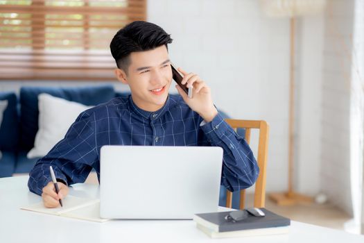 Young asian man talking phone and work from home with laptop computer, freelance male writing on notebook and speak on smartphone, stay home, domestic life, business and communication concept.