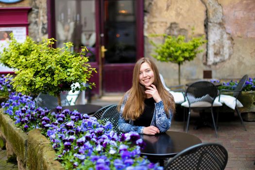 Young woman sitting at street cafe near blue flowers and green plants. Concept of cozy place in open air and resting.