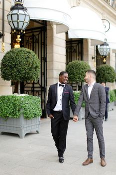 Afro american and caucasian handsome gays walking outside and holding hands in city. Concept of same sex male couple.