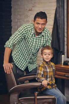 stylish little kid sitting on chair at barbershop with his young father on background, they are fooling around and having fun