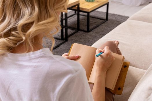 Close up woman hand writing on notebook. Young woman sitting on sofa in modern apartment, casual style indoor shoot