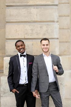 Afro american and caucasian happy smiling gays standing near building and wearing suits. Concept of lgbt and walking in city.