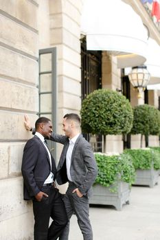 Afro american and european gays standing near building and wearing suits and talking outside. Concept of lgbt and walking in city.