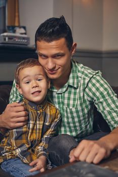 Young father and his stylish little son in the barbershop in the waiting room. They are waiting for the master, reading the fashion magazine and having a good time. Father hugs his son