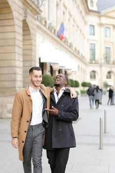 Caucasian handsome man in suit walking with afroamerican male person and hugging in city. Concept of happy gays and strolling.