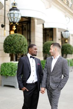 Afro american and caucasian gays walking and speaking outside in city. Concept of same sex male couple.