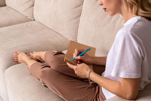 Close up woman hand writing on notebook. Young woman sitting on sofa in modern apartment, casual style indoor shoot