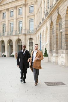 Caucasian man in suit running with afroamerican male person and holding hands in city. Concept of happy gays and strolling.