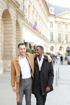 Caucasian happy man in suit walking with afroamerican male person and hugging in city. Concept of gays and strolling.