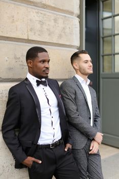 Afro american and caucasian handsome gays standing near building and wearing suits. Concept of lgbt and walking in city.