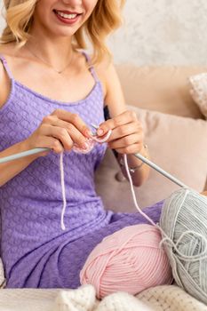 Close-up of woman in a lilac home dress knits a light pink scarf or plaid from natural threads on the bed. Close-up horizontal photo. Freelance creative working and living concept.