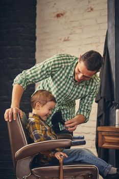 stylish little kid sitting on chair at barbershop with his young father on background, they are fooling around and having fun