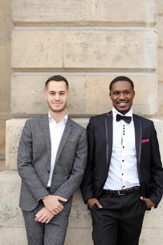 Afro american and caucasian gays standing near building and wearing suits. Concept of lgbt and walking in city.