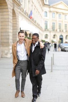 Caucasian smiling man in suit walking with afroamerican male person and hugging in city. Concept of happy gays and strolling.