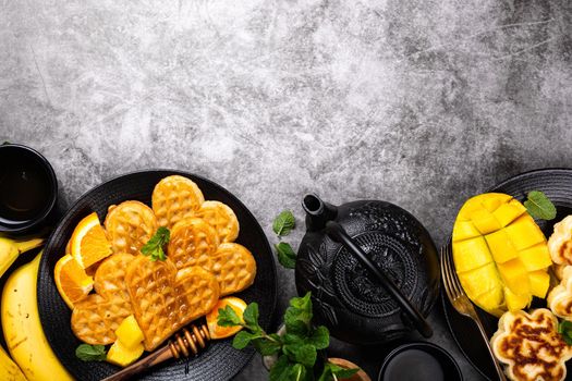 Healthy breakfast background with fresh hot waffles hearts, pancakes flowers with berry honey and exotic fruits over gray, top view, flat lay. Healthy food concept with copy space.