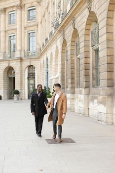 Caucasian handsome man running with afroamerican male person and holding hands in city. Concept of happy gays and strolling.