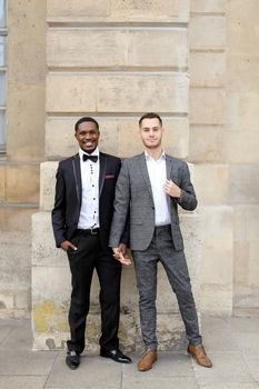 Afro american and european gays standing near building and wearing suits. Concept of lgbt and walking in city.