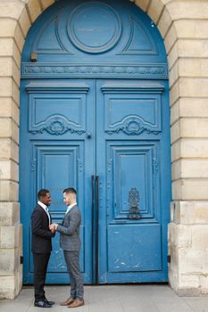 Afro american smiling man holding hands of caucasian boy in blue door background, wearing suit. Concept of same sex couple and gays.