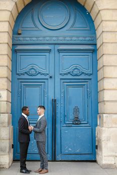 Afro american smiling man holding hands of caucasian guy in blue door background, wearing suit. Concept of same sex couple and gays.