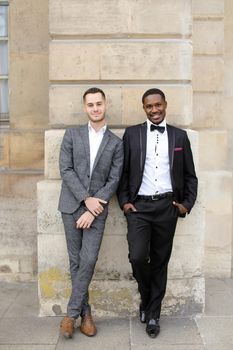 Afro american and caucasian happy gays standing near building and wearing suits. Concept of lgbt and walking in city.