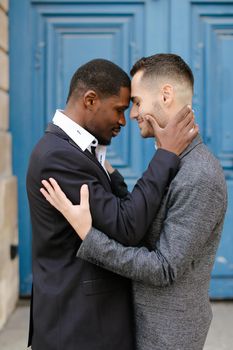 Afro american smiling man hugging caucasian guy in door background, wearing suit. Concept of same sex couple and gays.