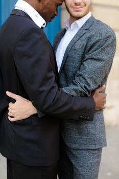 Afro american happy man hugging caucasian guy in door background, wearing suit. Concept of same sex couple and gays.