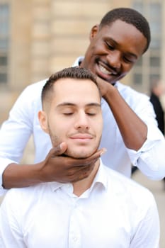 Afro american boy holding caucasian guy head by hands, wearing white shirt. Concept of stylish boy and haircut.