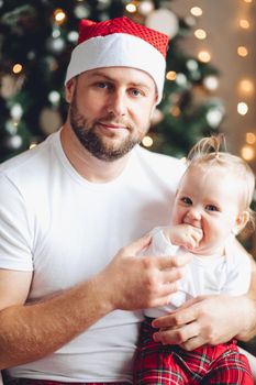 Portrait of a loving bearded dad in red Santa hat holding cute little daughter in arms.