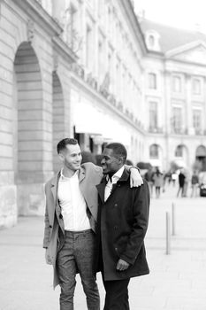 Caucasian man in suit walking with afroamerican male person and hugging in city. Concept of happy gays and strolling.