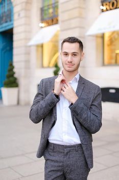 Young cacuasian male person wearing grey suit standing near building outdoors. Concept of fashion and businessman, urban life.