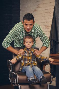 stylish little kid sitting on chair at barbershop with his young father on background, they are fooling around and having fun