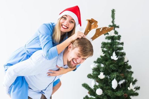 Funny man giving piggyback to his wife while they wearing Santa hats for Christmas holidays at home