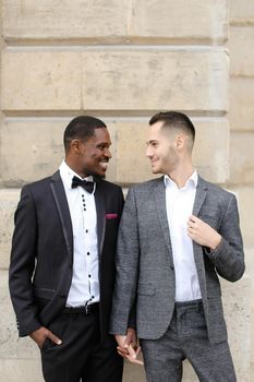 Afro american and caucasian smiling gays standing near building and wearing suits. Concept of lgbt and walking in city.