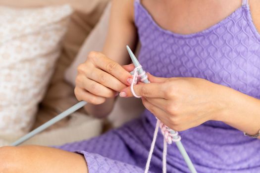 Close-up of woman in a lilac home dress knits a light pink scarf or plaid from natural threads on the bed. Close-up horizontal photo. Freelance creative working and living concept.