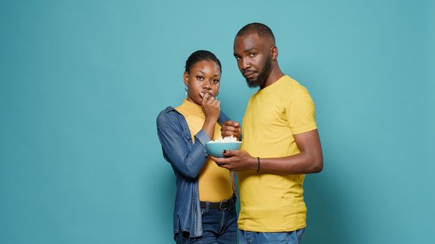 Couple eating popcorn snack from bowl in front of studio camera. Man and woman enjoying movie and watching television, standing over isolated background. People looking at show series.