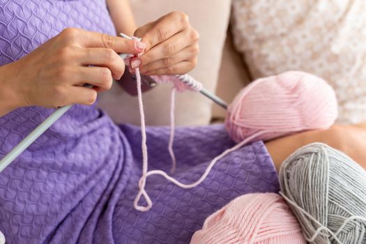 Close-up of woman in a lilac home dress knits a light pink scarf or plaid from natural threads on the bed. Close-up horizontal photo. Freelance creative working and living concept.