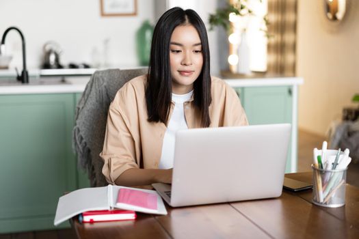 Asian girl studying at home, distance education concept. Female student learning with laptop. Young woman working remote, sitting in kitchen with books and computer.