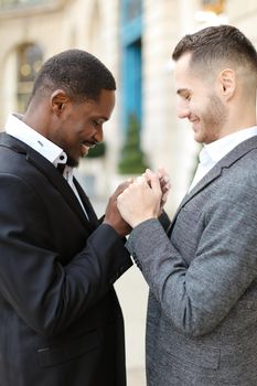 Afro american gay holding hands of caucasian boy outside, wearing suits. Concept of lgbt and same sex couple.