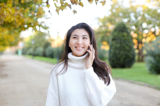 Asian smling woman wearing white sweater speaking by by smartphone walking in autumn park. Concept of chinese girl and modern technology.