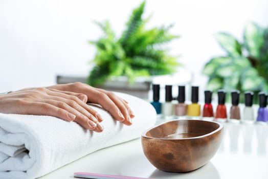 Female hands lying on white towel. Manicurist workspace with colorful nail polish bottles and wooden bowl with water. Female hand preparing for manicure. Professional nail care and beautician service.