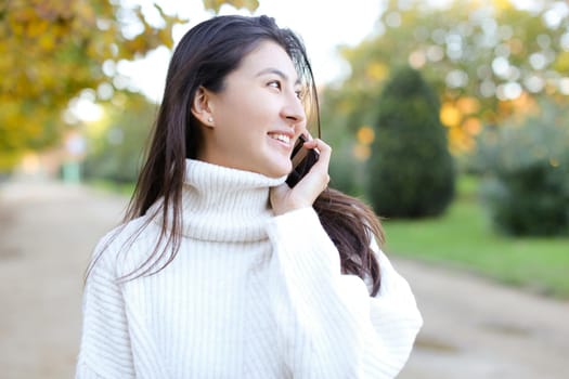 Asian happy woman wearing white sweater speaking by smartphone walking in autumn park. Concept of chinese girl and modern technology.