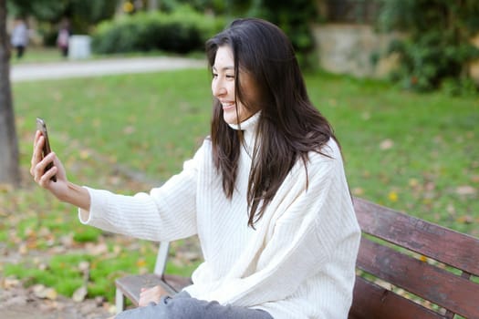 Japanese woman sitting on bench and making video call by smartphone. Concept of selfie and modern technology.