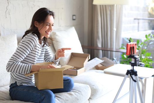 Young beautiful female blogger recording video while unpacking parcel at home