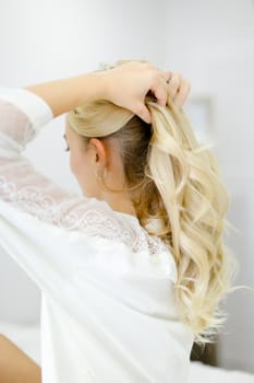 Young blonde caucasian woman wearing white closes touching curly hair. Concept of morning preparations and hairdo.