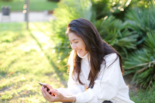 Asian female student sitting in park and typing by smartphone. Concept of modern technology and social networks.