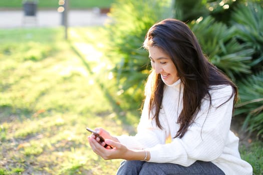 Asian female student white sweater sitting in park and typing by smartphone. Concept of modern technology and social networks.